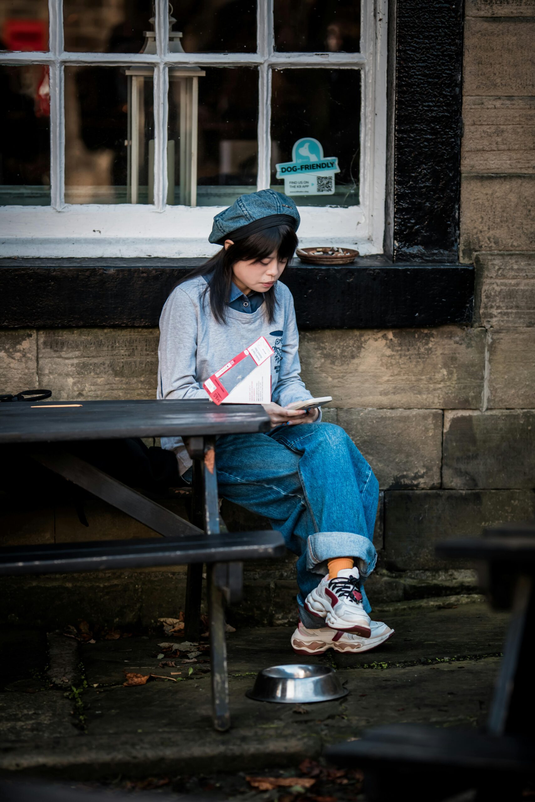 A young woman reads a book outside a cafe, dressed casually in denim.